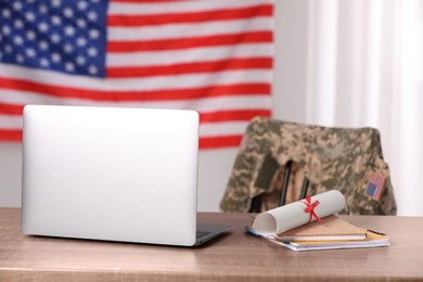 Laptop, notebooks and diploma on wooden table, chair with soldier uniform against flag of United States indoors. Military education