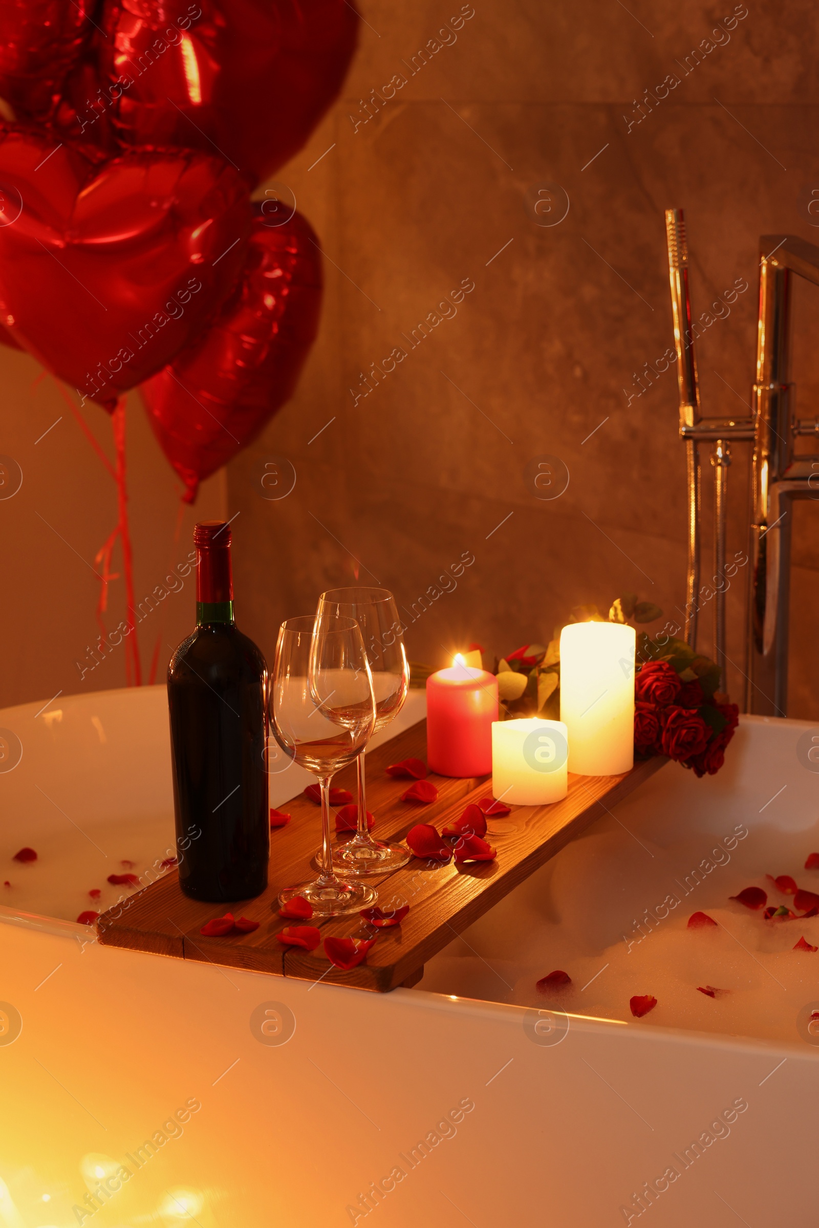 Photo of Wooden tray with wine, burning candles and rose petals on tub in bathroom. Valentine's day celebration