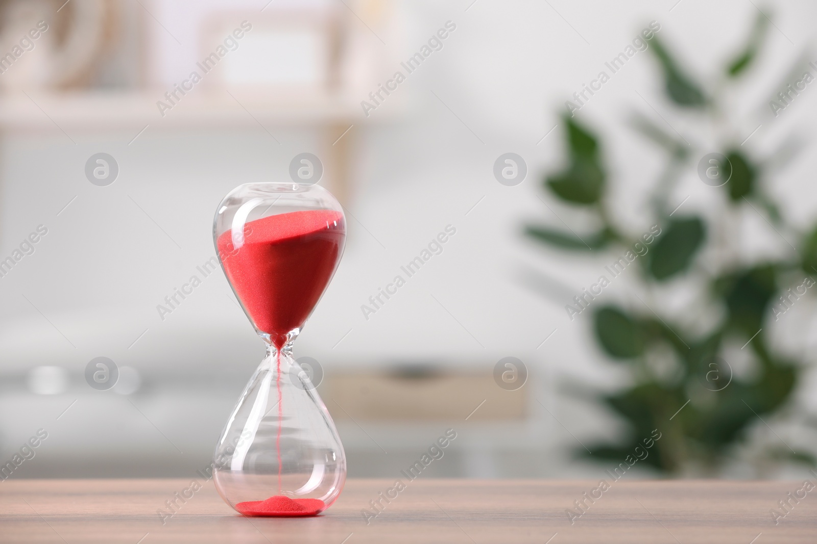 Photo of Hourglass with red flowing sand on table against blurred background. Space for text