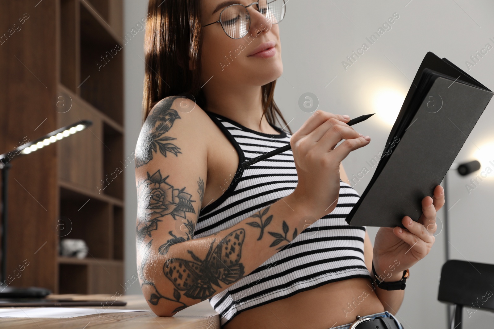 Photo of Beautiful woman with tattoos on arm drawing in sketchbook indoors