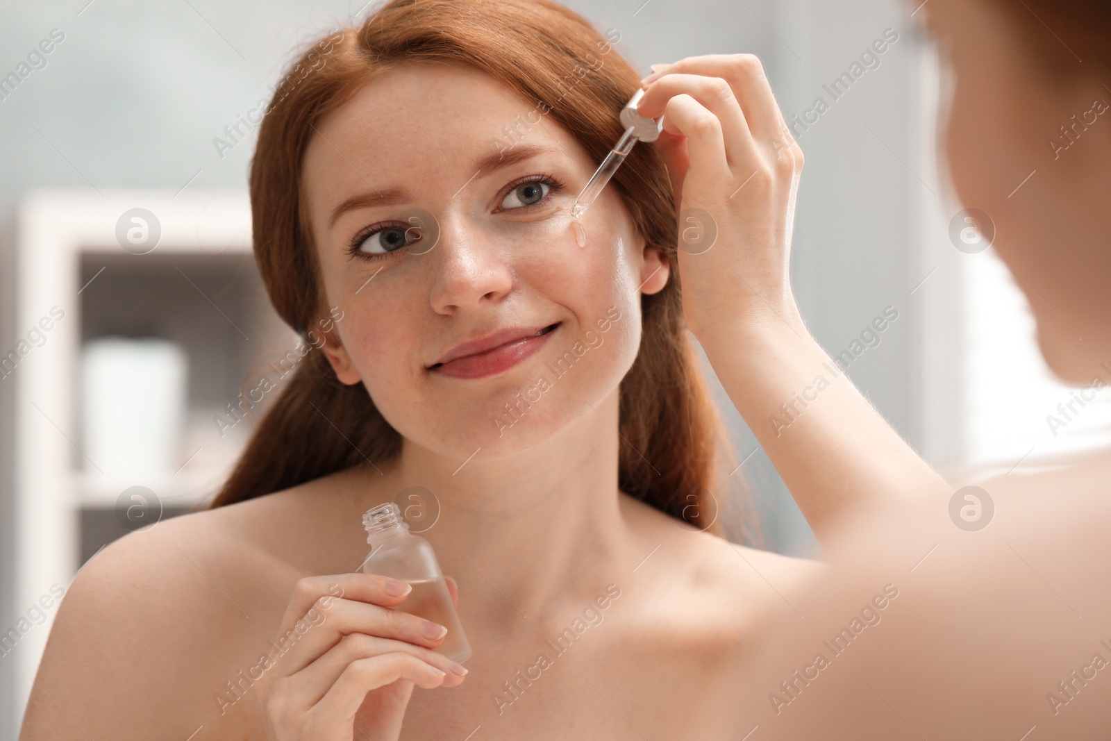 Photo of Beautiful woman with freckles applying cosmetic serum onto her face near mirror in bathroom