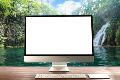 Modern computer with blank screen on white table and view of beautiful waterfall and lake