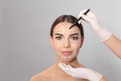 Doctor drawing marks on woman's face for cosmetic surgery operation against grey background