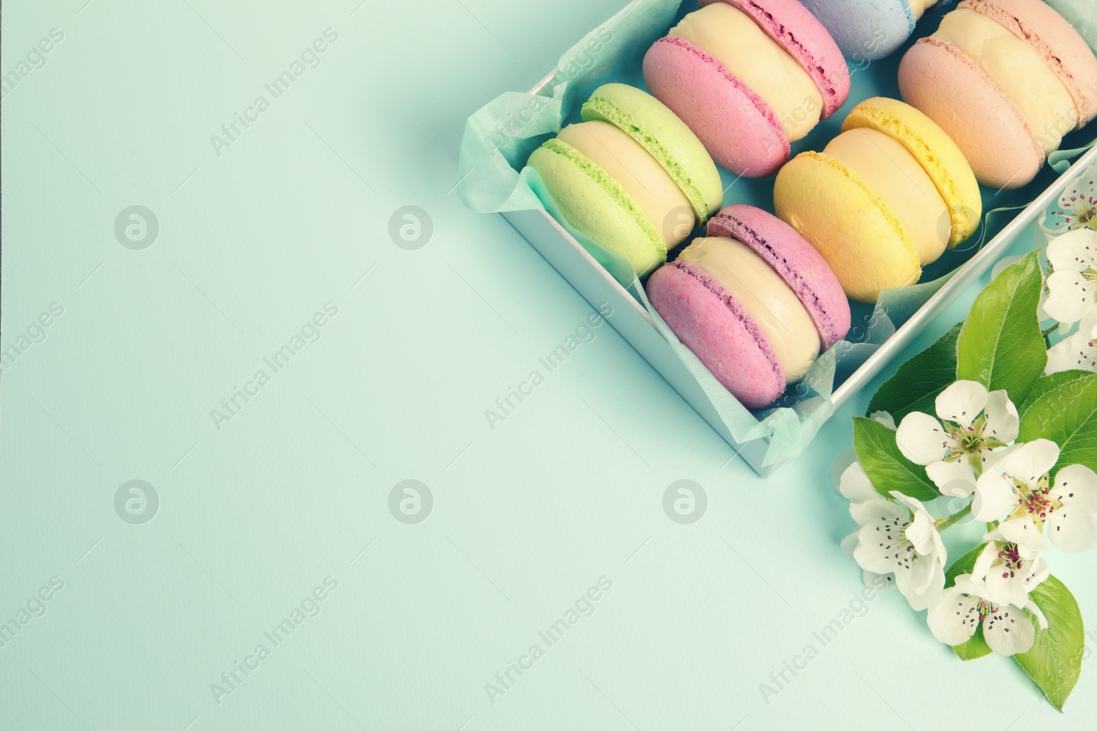 Photo of Many delicious colorful macarons in box and flowers on light blue background, flat lay. Space for text