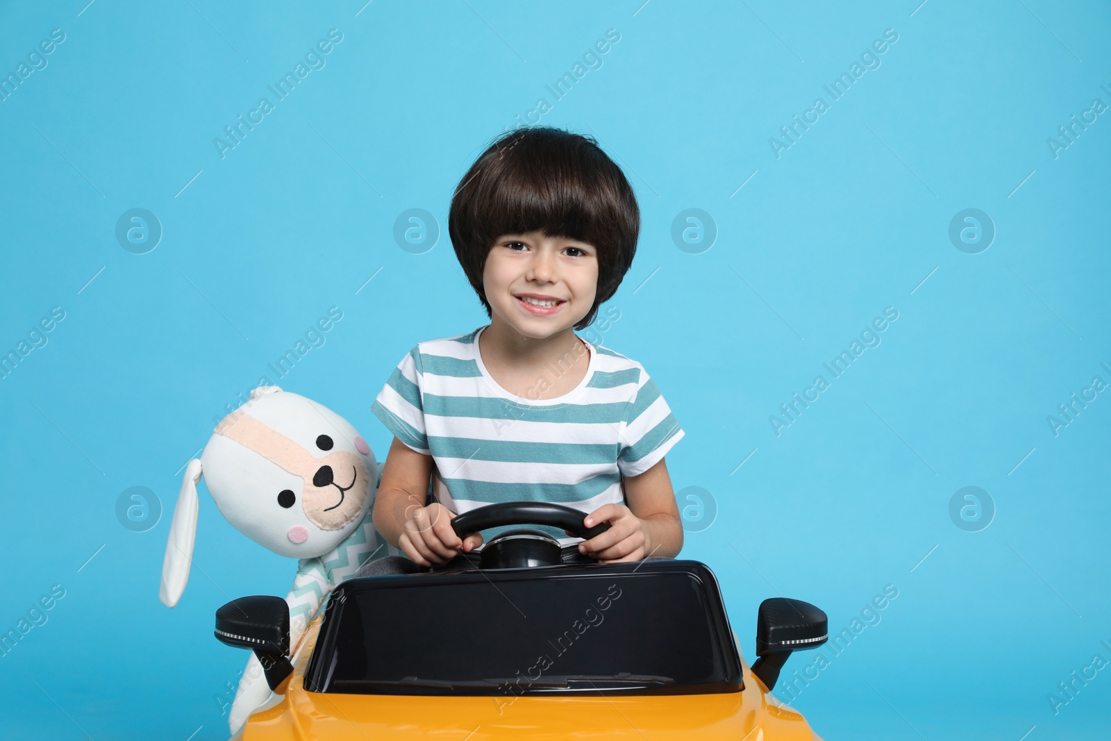 Photo of Cute little boy with toy bunny driving children's car on light blue background