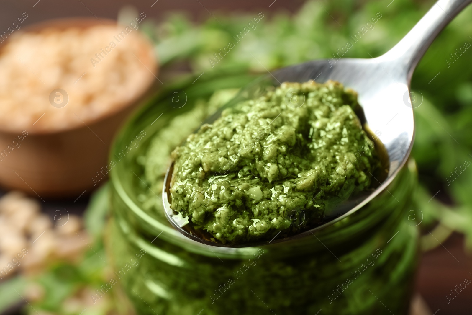 Photo of Spoon of tasty arugula pesto near jar with sauce, closeup