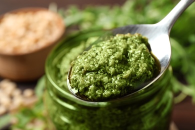 Photo of Spoon of tasty arugula pesto near jar with sauce, closeup