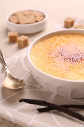 Photo of Delicious creme brulee in bowl, vanilla pods and spoon on table, closeup