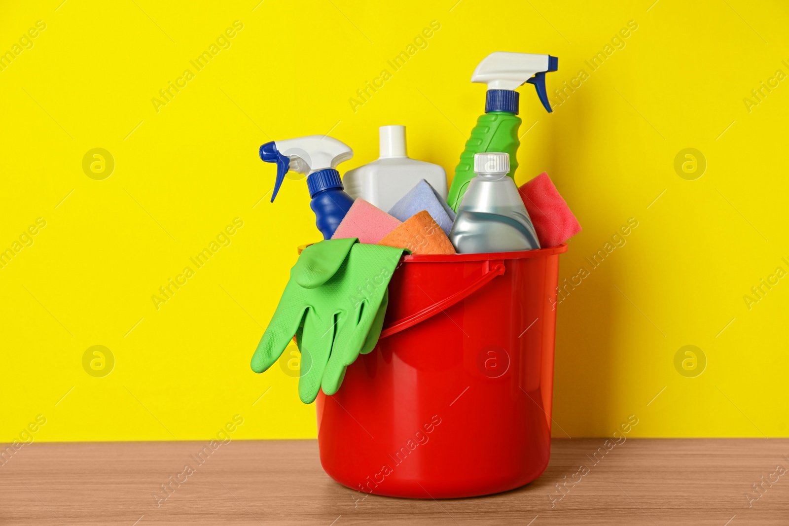 Photo of Bucket with different cleaning supplies on wooden floor near yellow wall