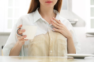 Woman with spilled coffee over her shirt at table in kitchen, closeup