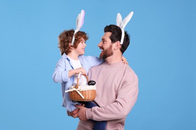 Photo of Happy father and son in cute bunny ears headbands on light blue background. Boy holding Easter basket with painted eggs