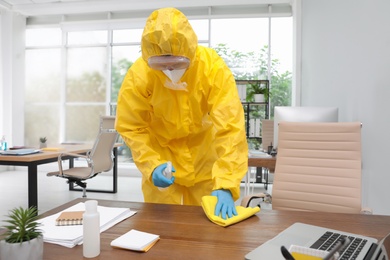 Photo of Janitor in protective suit disinfecting office furniture to prevent spreading of COVID-19