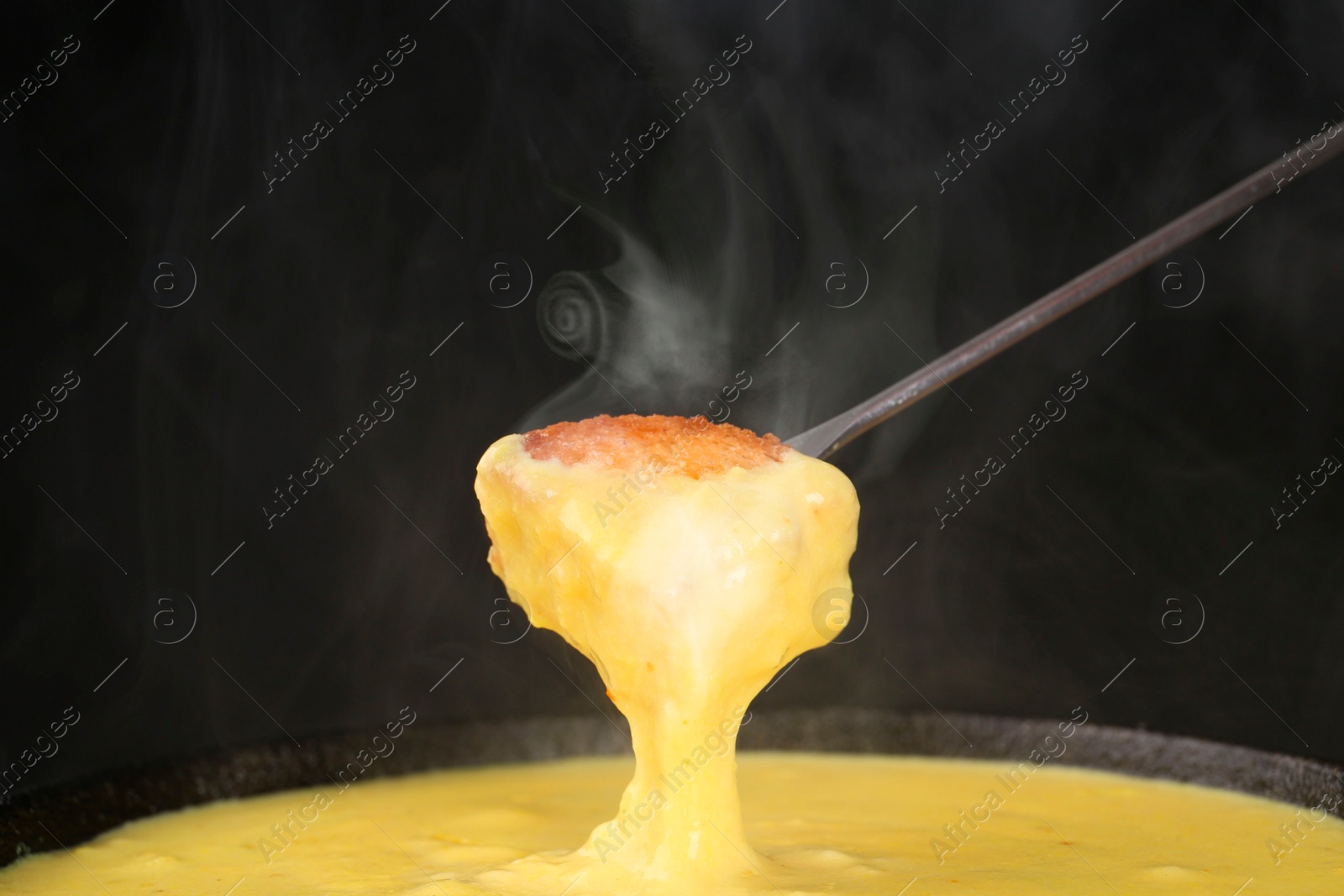 Image of Dipping piece of bread into fondue pot with tasty melted cheese against dark background, closeup