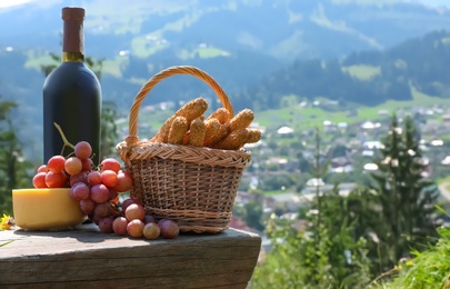 Photo of Bottle of red wine with ripe juicy grapes and other food for picnic on bench against mountain landscape