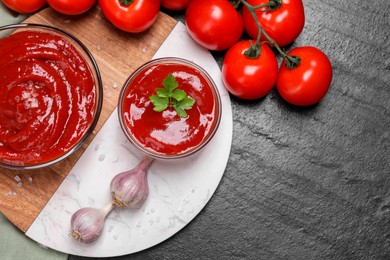Photo of Organic ketchup in bowls, fresh tomatoes and garlic on black table, flat lay with space for text. Tomato sauce