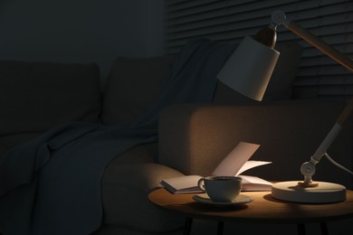 Stylish lamp, book and cup of tea on side table near sofa indoors