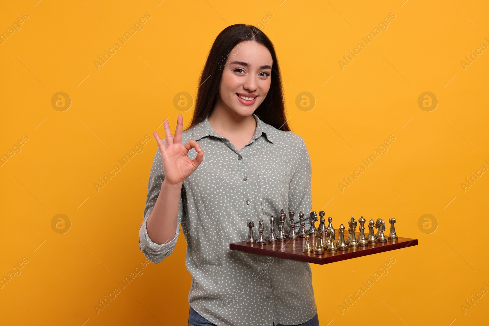 Photo of Happy woman holding chessboard with game pieces and showing OK gesture on orange background