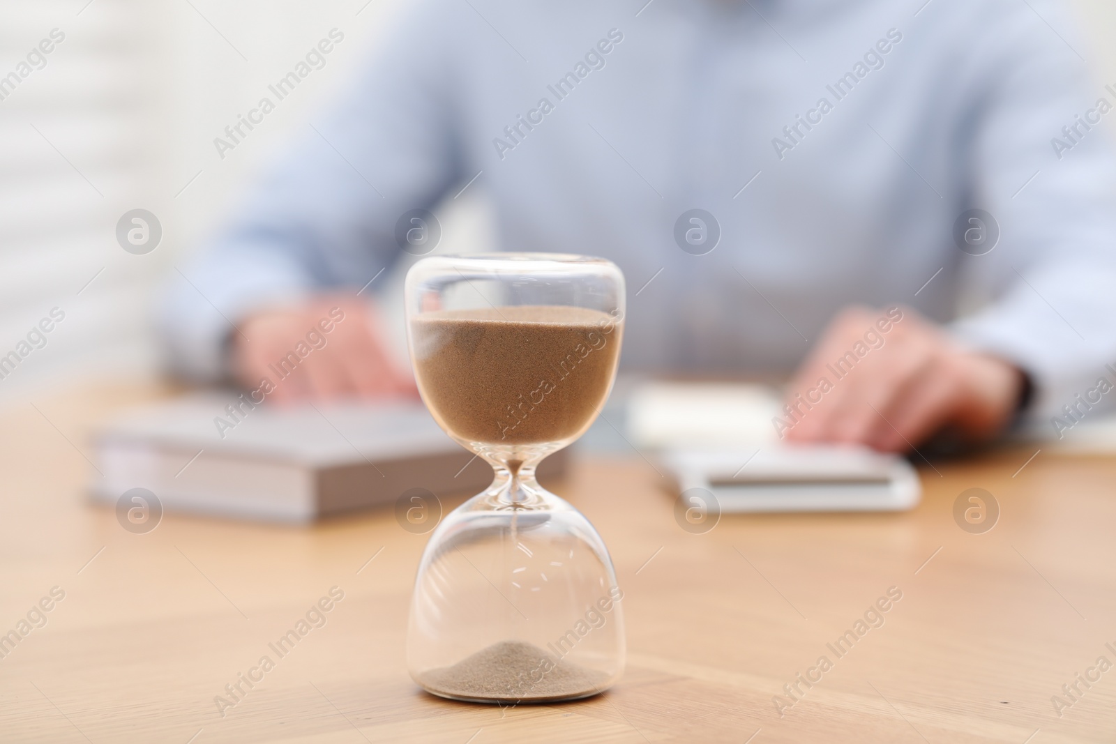 Photo of Hourglass with flowing sand on desk. Man using calculator indoors, selective focus