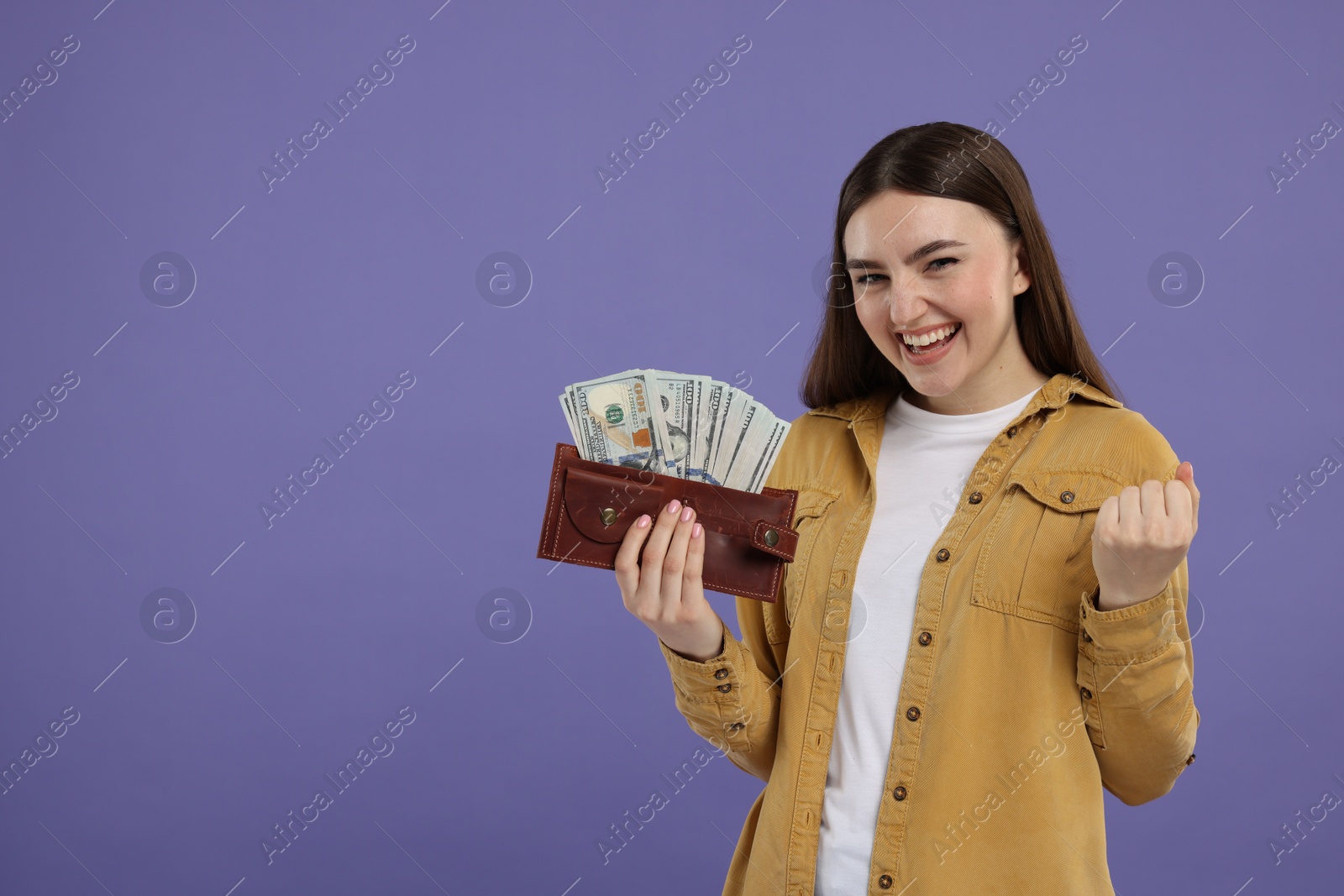 Photo of Excited woman holding wallet with dollar banknotes on purple background, space for text