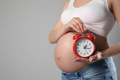 Young pregnant woman holding alarm clock near her belly on grey background, closeup and space for text. Time to give birth