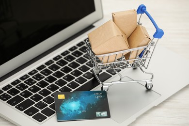 Photo of Online payment concept. Small shopping cart with bank card, boxes and laptop on white table, closeup