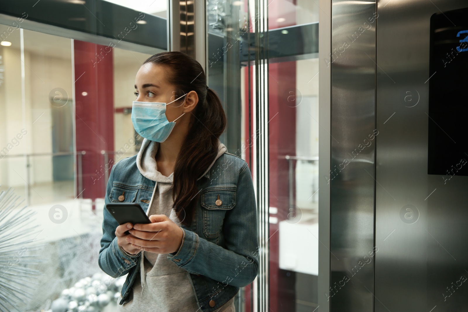 Photo of Young woman with disposable mask and smartphone in elevator. Dangerous virus