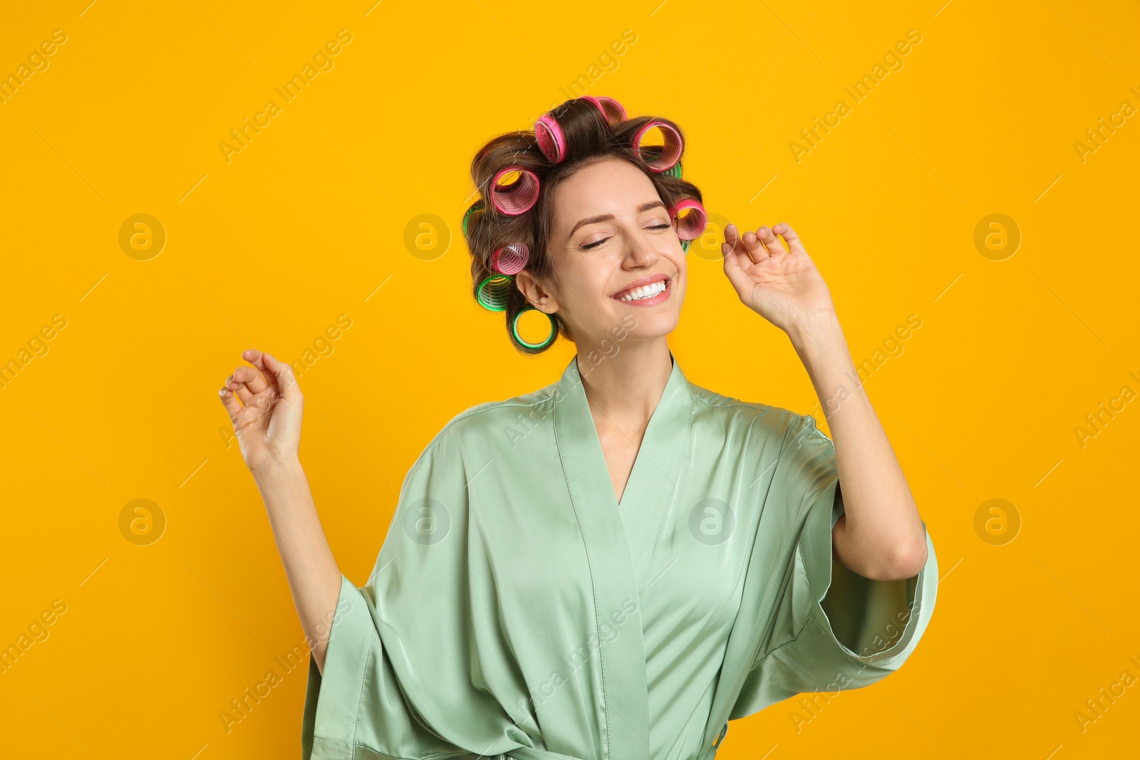 Photo of Happy young woman in silk bathrobe with hair curlers dancing on yellow background