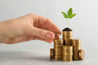 Photo of Woman putting coin onto stack with green plant on white marble table, closeup. Investment concept