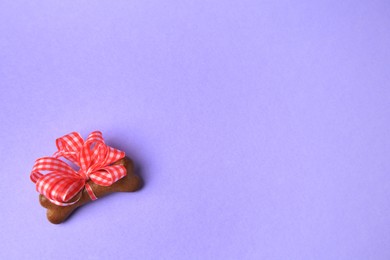 Photo of Bone shaped dog cookie with red bow on purple background, above view. Space for text