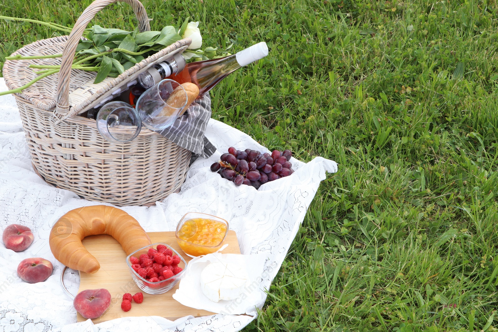 Photo of Picnic basket with tasty food, flowers and cider on blanket outdoors