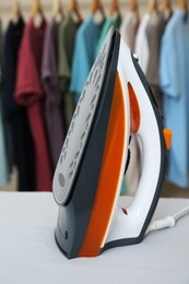 Photo of Modern iron on light table near rack with colorful clothes, closeup