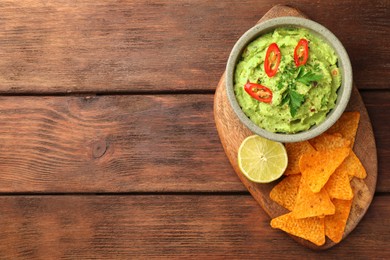 Photo of Bowl of delicious guacamole, lime and nachos chips on wooden table, top view. Space for text