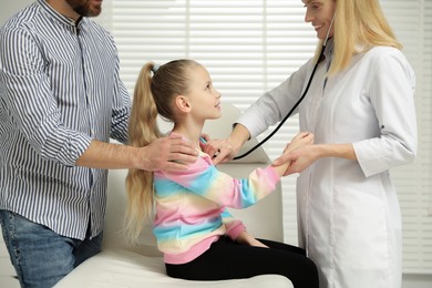 Photo of Father and daughter having appointment with doctor. Pediatrician examining little patient with stethoscope in clinic