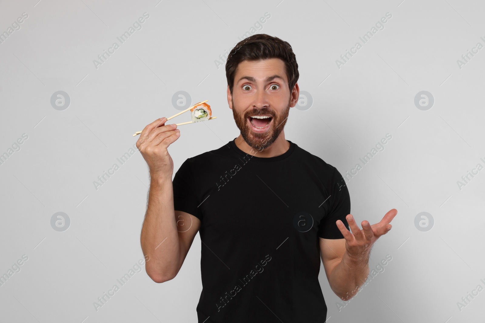 Photo of Emotional man holding sushi roll with chopsticks on light grey background