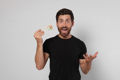 Emotional man holding sushi roll with chopsticks on light grey background