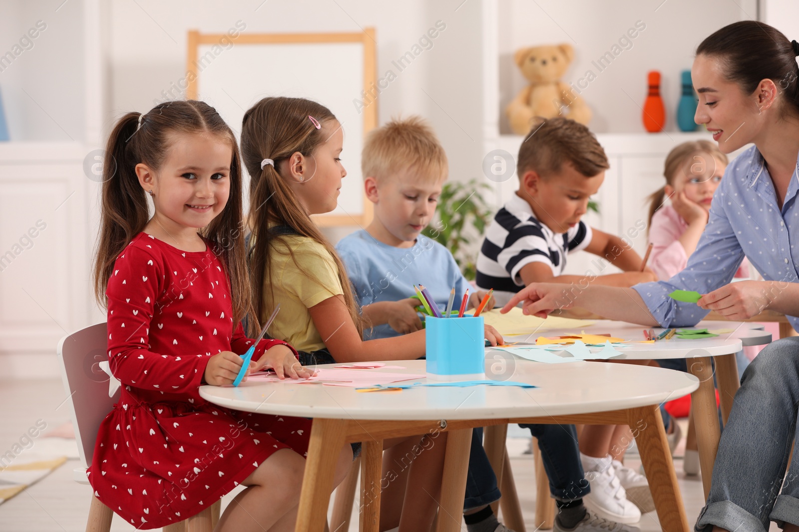 Photo of Nursery teacher and group of cute little children making toys from color paper at desks in kindergarten. Playtime activities