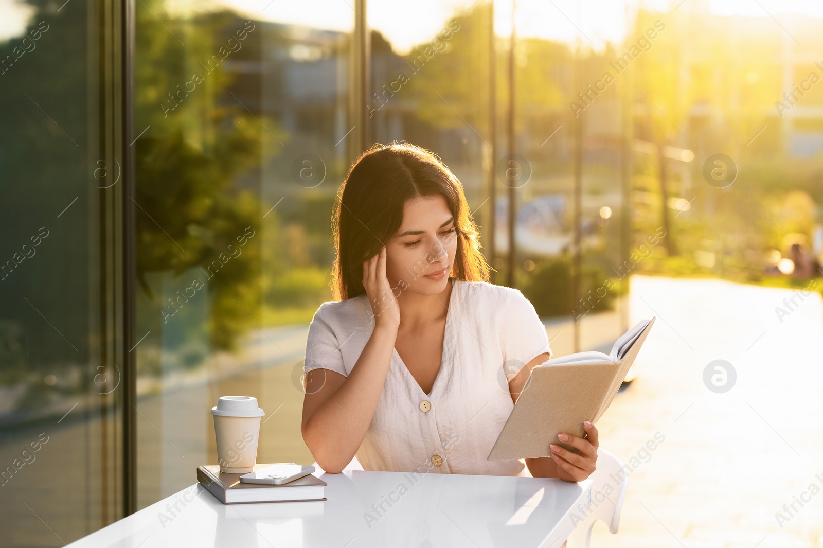 Photo of Young woman with coffee reading book in outdoor cafe on sunny day