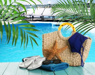Image of Stylish bag with beach accessories on light blue wooden surface near outdoor swimming pool