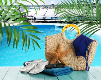 Stylish bag with beach accessories on light blue wooden surface near outdoor swimming pool