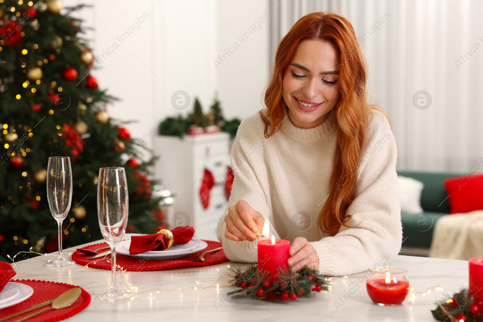 Photo of Beautiful young woman lighting candle at table in room decorated for Christmas