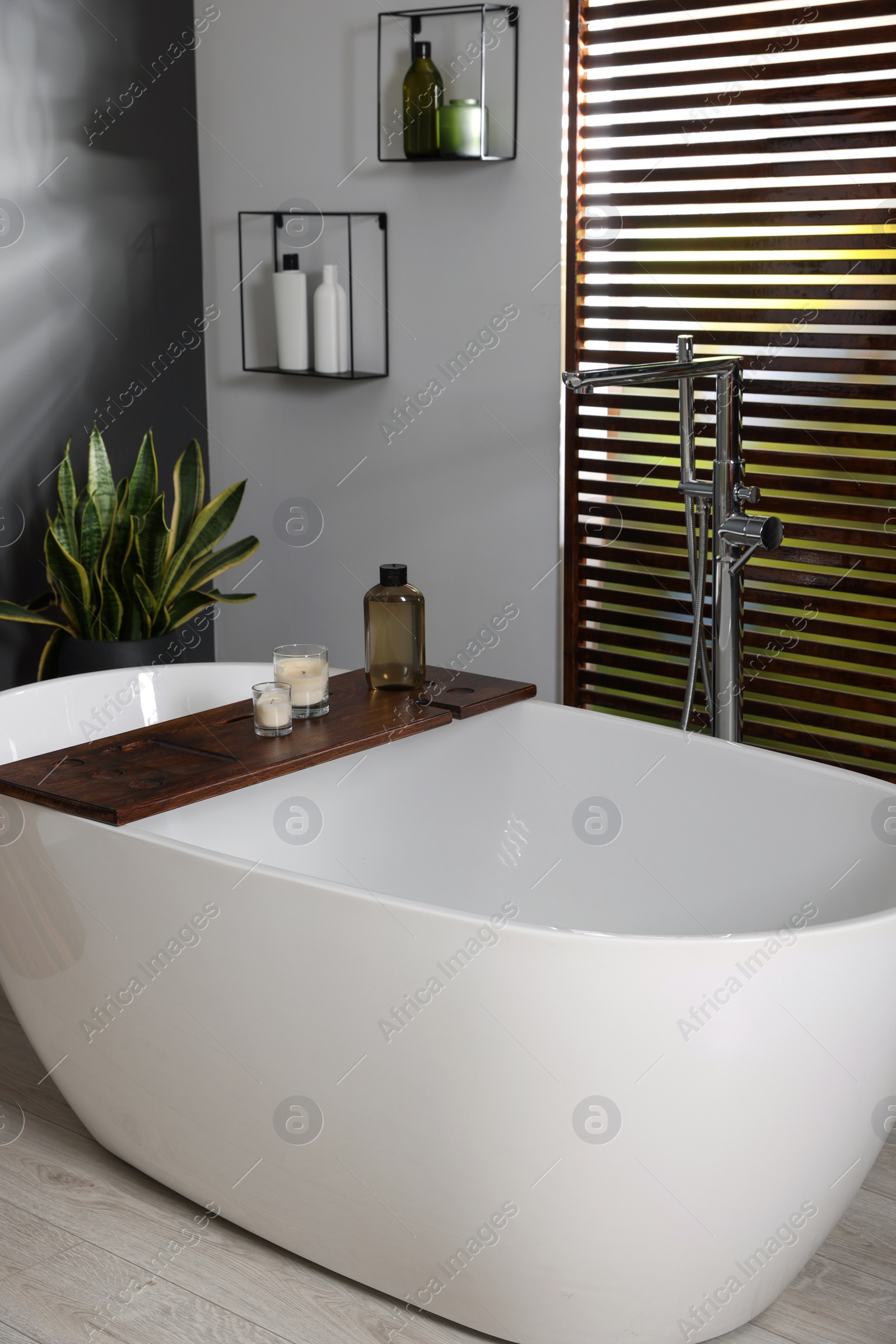 Photo of Stylish bathroom interior with ceramic tub, candles and care products on wooden bath tray