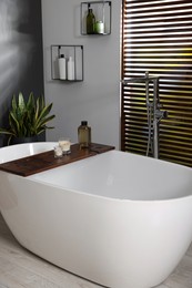 Stylish bathroom interior with ceramic tub, candles and care products on wooden bath tray