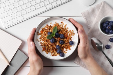 Woman holding bowl of tasty granola with blueberries at white wooden table with computer keyboard, top view