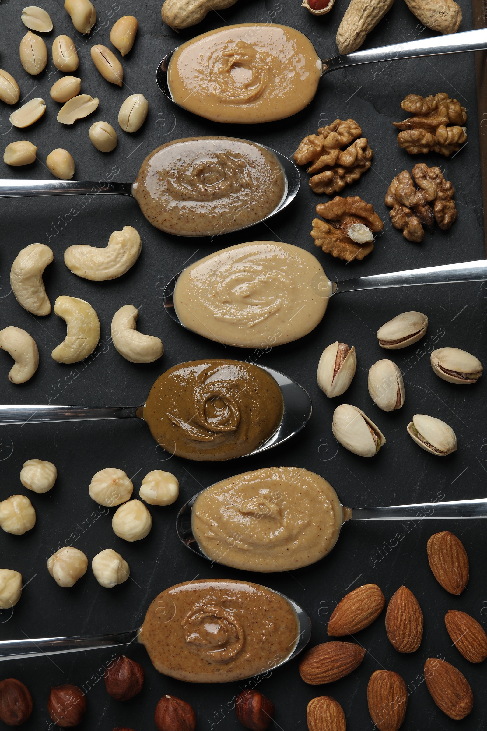 Photo of Tasty nut butters in spoons and raw nuts on dark gray table, flat lay