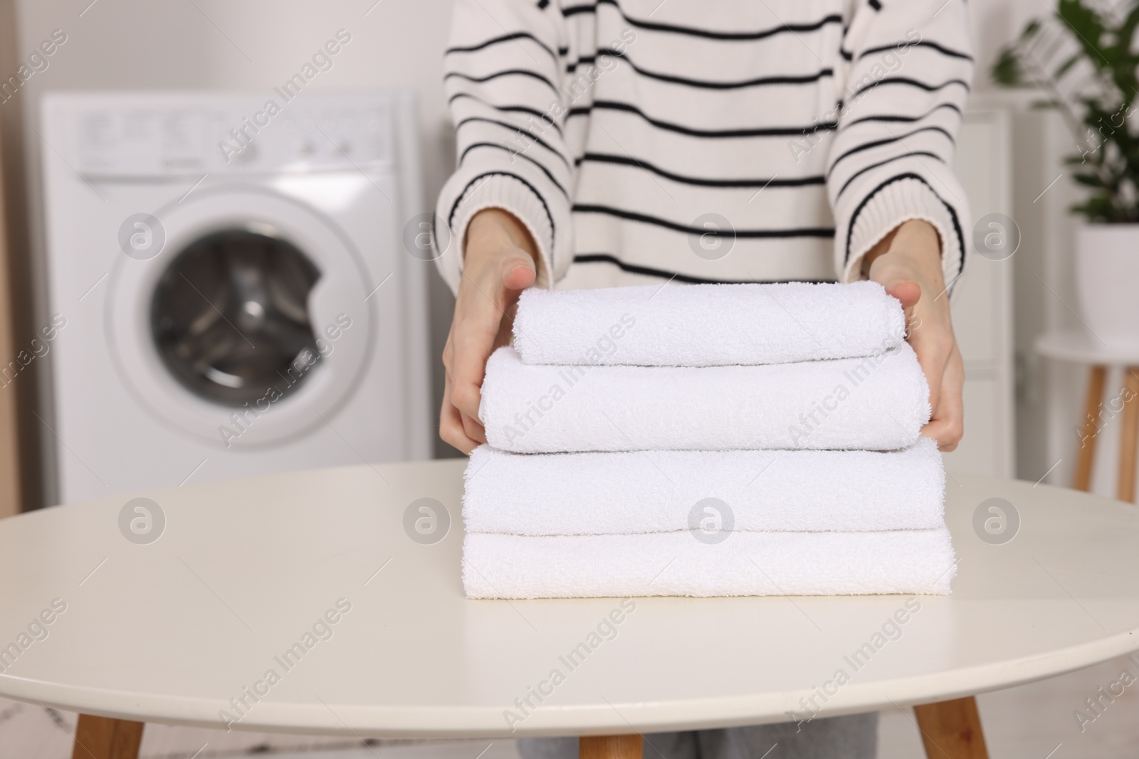 Photo of Woman with stack of folded towels at white table in laundry room, closeup. Space for text