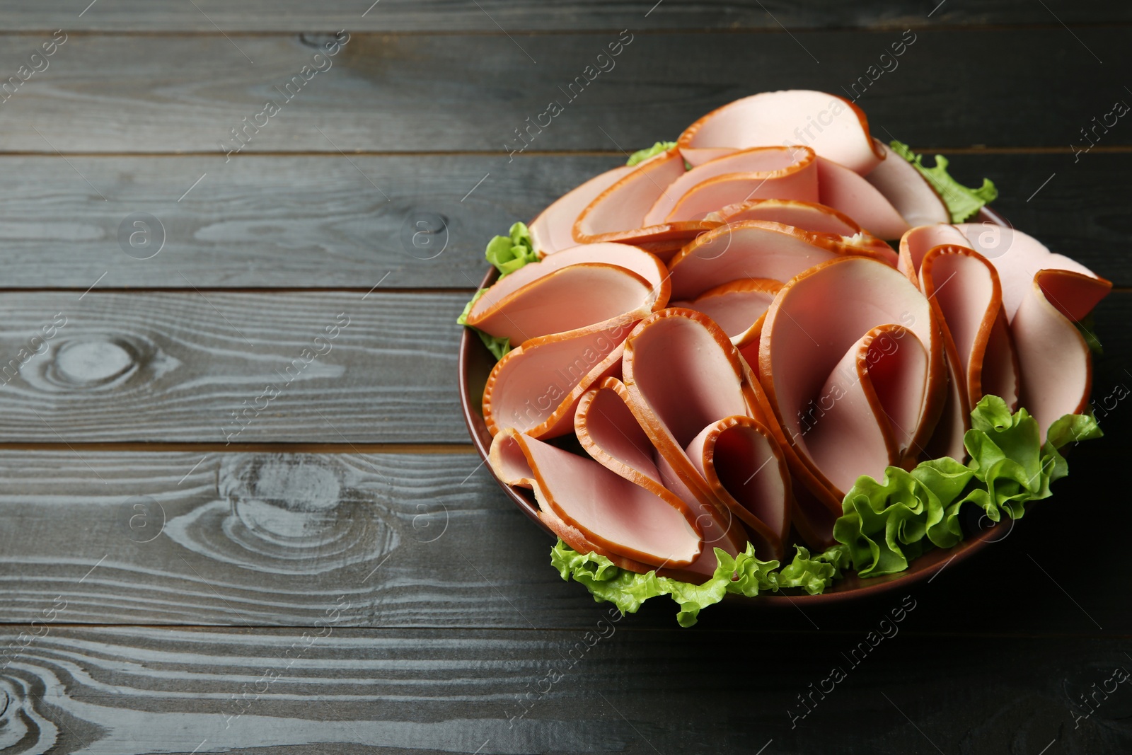 Photo of Slices of delicious boiled sausage with lettuce on dark wooden table, space for text