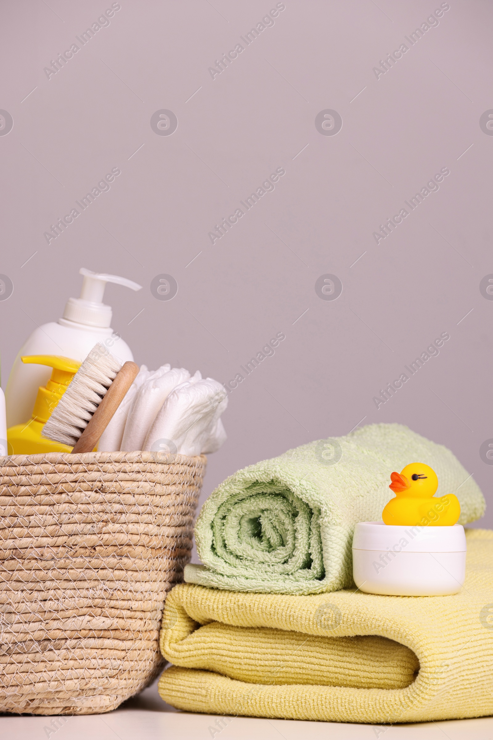 Photo of Wicker basket with baby cosmetic products, bath accessories and rubber duck on white table against grey background