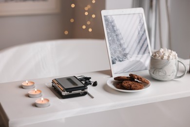 Photo of White wooden tray with tablet, cassette player, cookies and burning candles on bathtub in bathroom