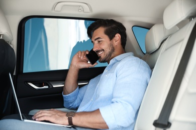 Photo of Attractive young man working with laptop and talking on phone in luxury car