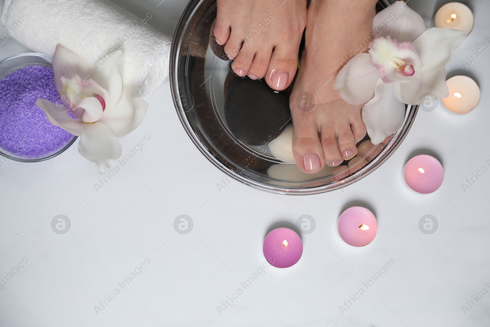 Photo of Woman soaking her feet in bowl with water, flower and spa stones on white marble floor, top view. Pedicure procedure